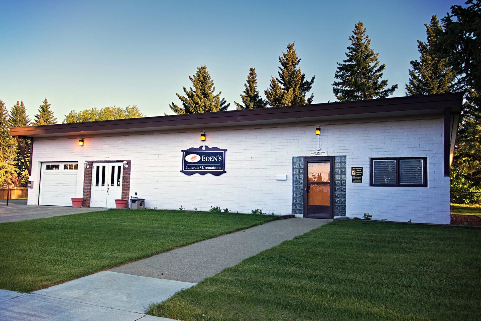The front of Eden's Funeral Home in Pincher Creek at dusk: white brick, a chapel entrance with double doors beside a garage bay, a separate family entrance with glass-block surround, and an Eden's Funerals & Cremations sign between them.
